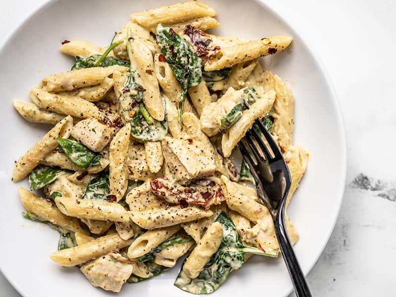 This super lush and Creamy Pesto Chicken Pasta is perfect for busy weeknights. Everything cooks in one pot and is done in under 30 minutes! BudgetBytes.com Close up of one pot creamy pesto chicken pasta in a shallow bowl with a black fork.