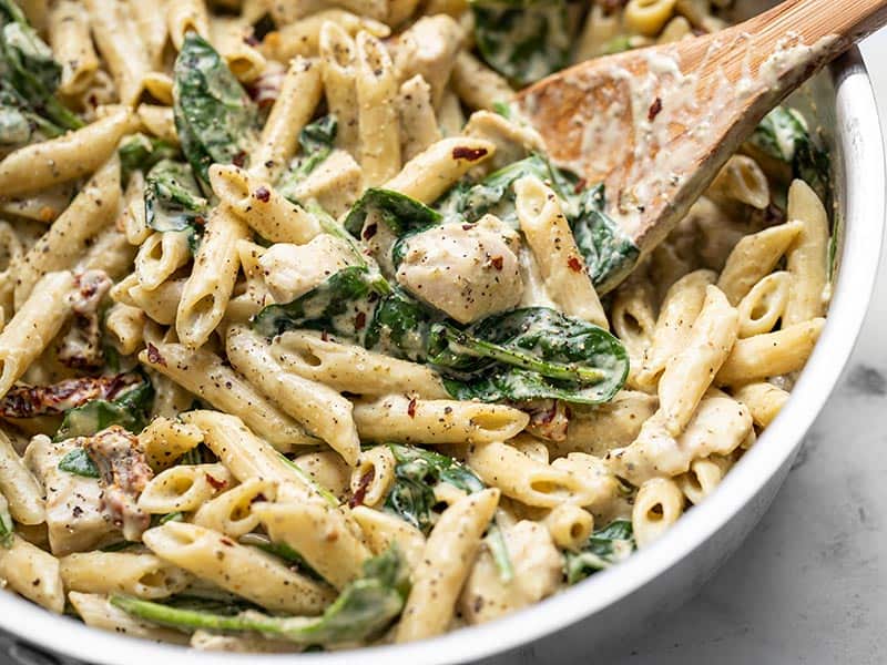 This super lush and Creamy Pesto Chicken Pasta is perfect for busy weeknights. Everything cooks in one pot and is done in under 30 minutes! BudgetBytes.com Close up of One Pot Creamy Pesto Chicken Pasta in the skillet with a wooden spoon.