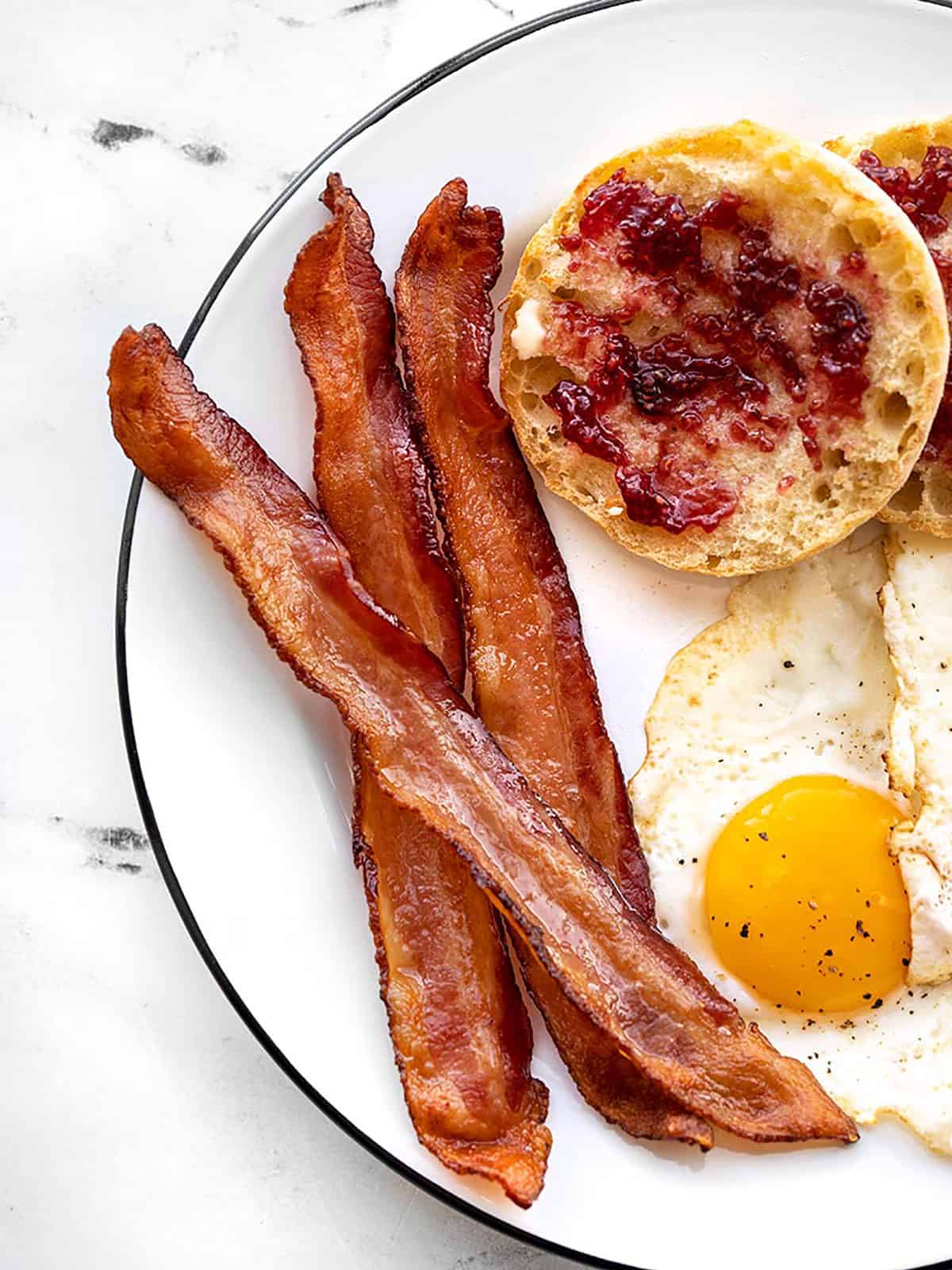 Learn how to cook bacon in the oven with very little effort and no hot splattering bacon grease! Perfect, crispy bacon in the oven every time. BudgetBytes.com Close up of crispy oven baked bacon on a plate with a fried egg and English muffin.