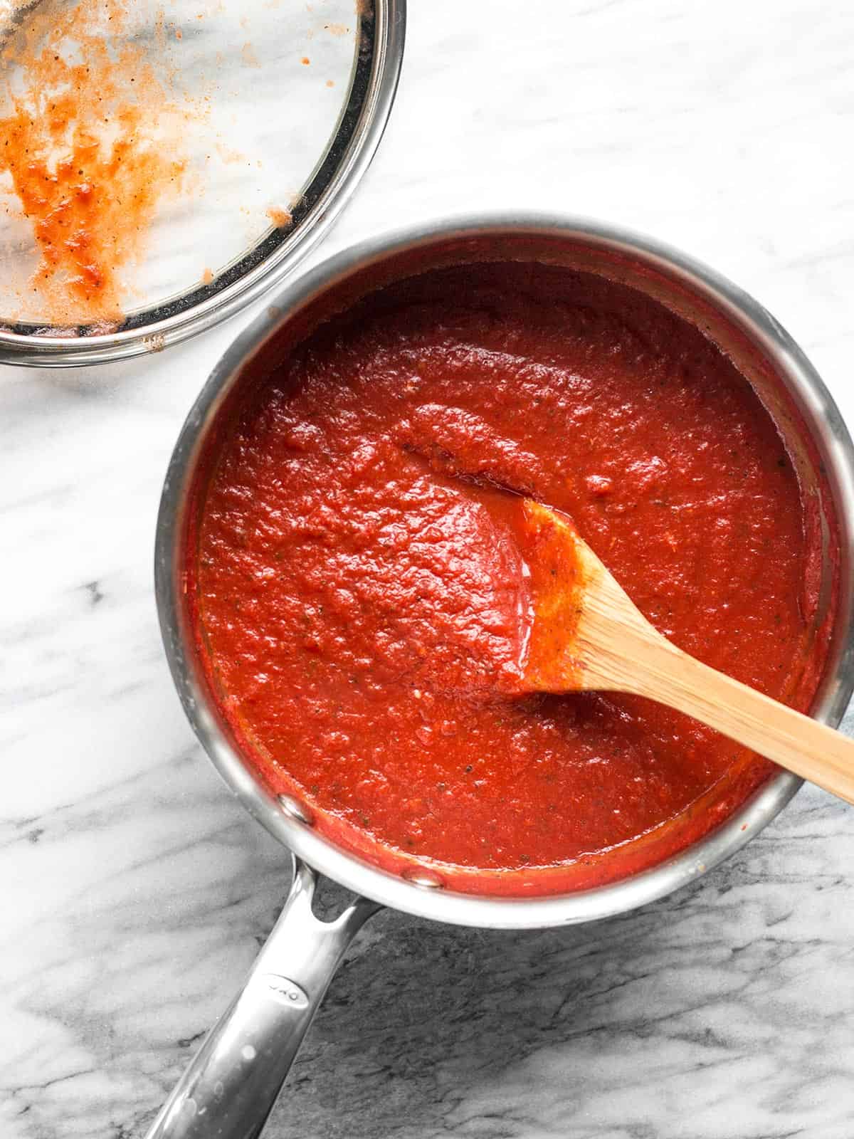 Use this Thick and Rich Pizza Sauce Recipe to make your own sauce at home in minutes with just a few pantry staples. Use now, or freeze for later! BudgetBytes.com Overhead view of a sauce pot full of pizza sauce with a wooden spoon in the center.